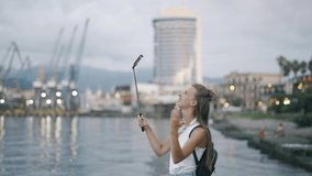 A young attractive woman blogger or vlogger is recording content for channel and looking at telephone and talking with beautiful city background. Cheerful girl is traveling and talking via video link.