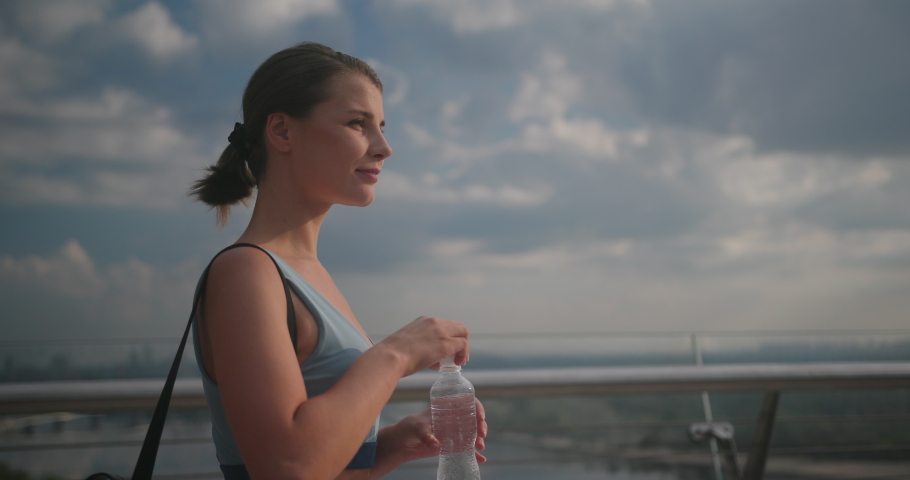 Young athletic woman drinks water on bright sunny day. Lady drinks water from transparent plastic bottle against blue sky with clouds slow motion Royalty-Free Stock Footage #1092315685