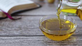 Hand holding glass jar and pouring out pure virgin olive oil into a container on a wooden table with an open Holy Bible Book, olives, and a green branch. Symbol of God's Holy Spirit. A closeup.