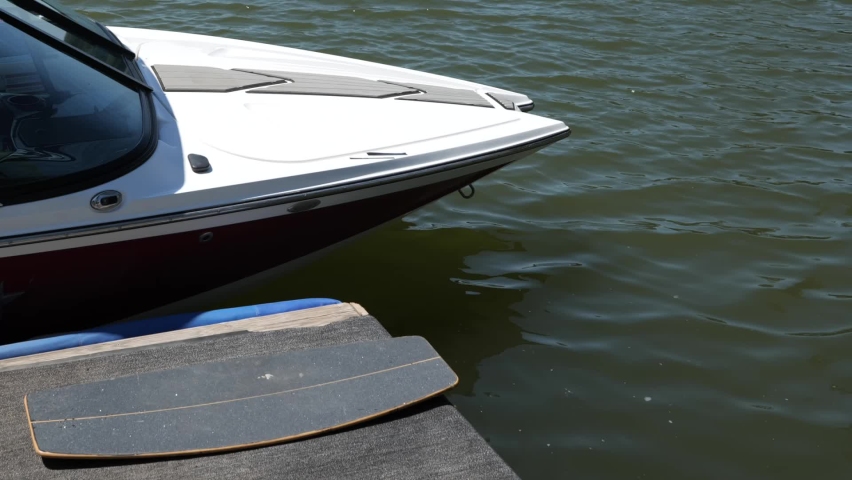 Wakeskate board lying next to a ski boat, wakeboard boat which is docked at the pier. A calm and empty lake in the background. Adventure, leisure, action sports scene.  Royalty-Free Stock Footage #1092326027