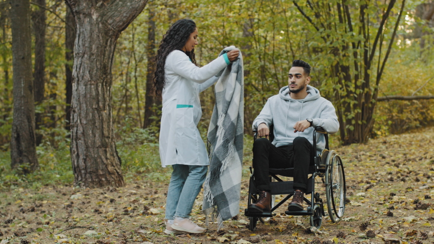 Young nurse in medical gown covers guy with warm blanket takes care patient in wheelchair caring girl visits friend in rehab walks in autumn hospital park enjoy walking outdoors rehabilitation center