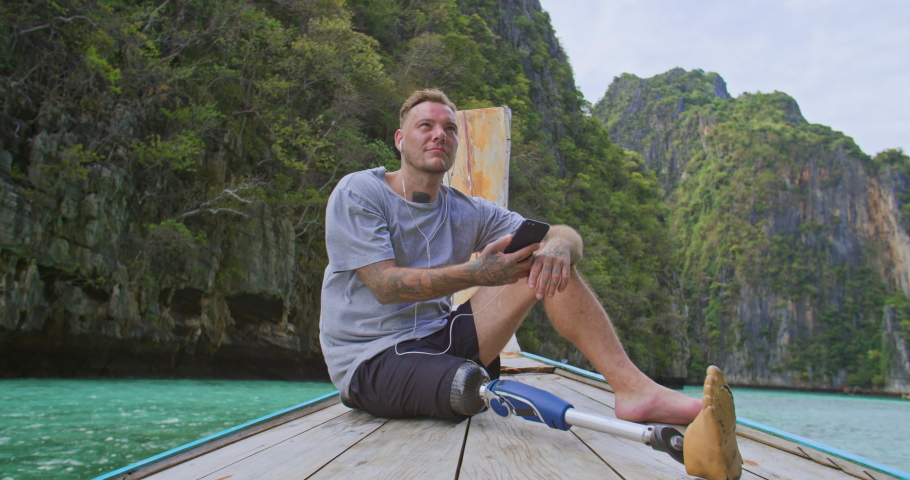 Man with artificial bionic leg prosthesis sitting on the front of long tail boat listening music while enjoying amazing beautiful seascape during summer vacation travel in Phi Phi Islands, Thailand.