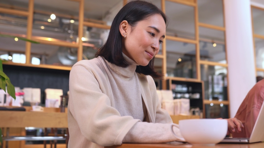 Happy smiling inspired young Asian ethnic woman enjoying morning coffee in cafe relaxing after remote work or learning, dreaming using laptop computer sitting at table in coffeeshop. Royalty-Free Stock Footage #1092331913