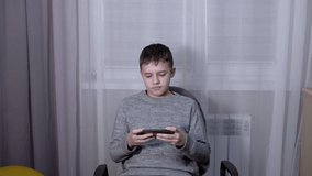 Handsome boy talking on a smartphone online in Room. A teenager communicates on a mobile phone with friends, and parents, sitting in an armchair. Child waving hello, goodbye. Internet. Home interior.