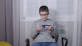 Smart Boy in Glasses Watching a Video in a Mobile App on a Smartphone. A teenager sits in a chair, writes messages, SMS in a chat. Online learning, video game viewing. Internet addiction. Lifestyle.