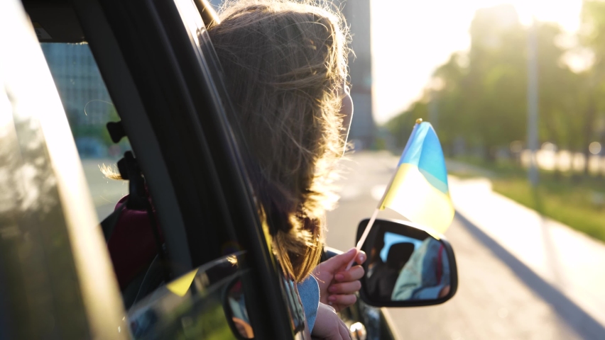 Caucasian girl in car window with Ukrainian flag in hand. Kid smiles at wind looking out vehicle window. Beautiful small female child holding of Ukraine out of an automobile. Family trip. Close up | Shutterstock HD Video #1092335291