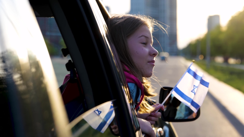 Joyful little girl holding Israeli flag in hand looking out of car window. Close up shot of small pretty child travels in Israel. Patriotic. Family trip. Travel concept. Slow motion Royalty-Free Stock Footage #1092335303