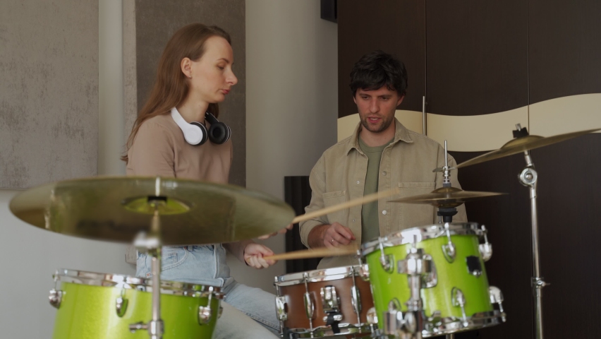 Young woman takes drum lesson from male teacher in professionally equipped music studio. Woman energetically plays drums and cymbals man watches performance