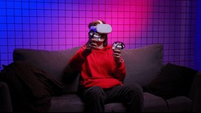 Girl in goggles uses finger-tracking controllers to play fun video game. Lady gamer in red hoodie sits on sofa in room with neon lights slow motion