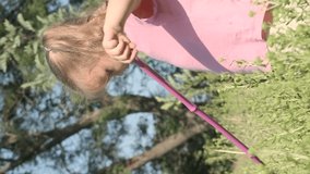 VERTICAL VIDEO: Little girl plays with butterfly net of tall grass in city park. Cute little girl is playing with aerial insect net in meadow on sun day. Slow motion