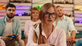 A close up of a blonde girl with long hair tied back and wearing black glasses is competing in a video game against her handsome friend with a beard