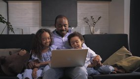 African man and children make a video call sitting at sofa.Black family looking at laptop in home interior.Smiling children watching TV shows with Father.