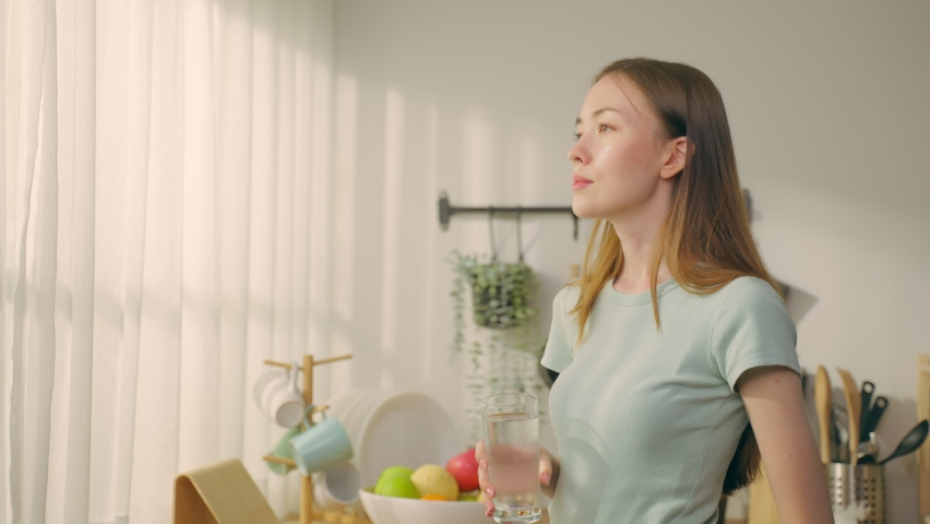 Beautiful Caucasian woman drinking a glass of water in kitchen at home.  Attractive young thirsty girl holding clean mineral natural in cup after waking up and sip in morning for health care in house. Royalty-Free Stock Footage #1092363463