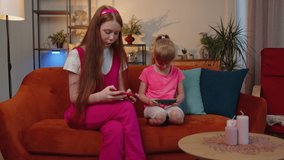 Worried funny girls friends siblings enthusiastically playing shooting video games on smartphone. Happy two female child kids family on couch at home, using smartphone gadget app with drive simulator