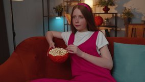 Excited young teen child girl sitting on sofa eating popcorn and watching interesting tv serial, sport game film, online social media movie content online at home. Kid enjoying domestic entertainment