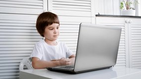 A focused and attentive child sits and does a task on a laptop. Close-up. 5 year old boy solves a test by a distance online education program. Smart kid. White room, furniture, t-shirt. Copy space.