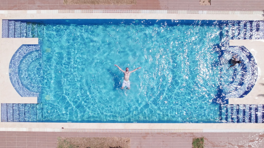 Top down aerial view of young man lies down backstroke on a rippling water surface in a swimming pool. Man relaxes at swimming pool lying on a back. High angle view