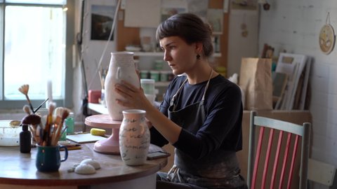 Creative woman performs individual order in pottery studio for production of clay pots, jag and other various things for home interior decor and gifts. Handmade vases for sale in craft store. 