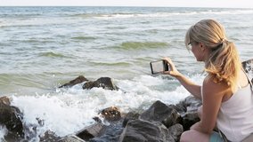 Beautiful happy young woman in mild light clothes with a careful braid, takes pictures of the landscape of the sea horizon on a modern phone while sitting on wet stones. HD slow-mo video