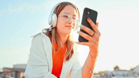 Happy teenage girl with headphones holds phone in her hands and watches video with smile. Using social networks and modern technologies for communication and entertainment. Summer leisure in nature.