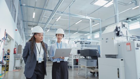 Two Female Heavy Industry Employees Wearing Hard Hats at Factory. Walking and Discussing Industrial Machine Facility, Using Laptop Computer. Asian Engineer and African American Technician at Work.