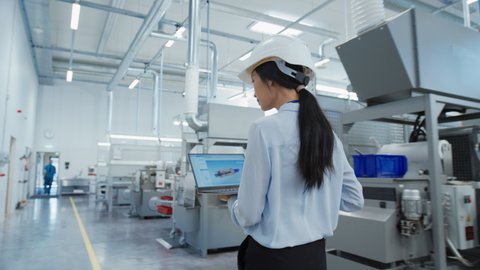 Research and Development Specialist in a Factory Facility, Holding Laptop Computer and Walking in Heavy Industry Machine. Asian Quality Control Manager Investigating the Work Space.
