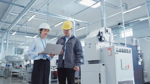 Portrait of Two Young Asian Heavy Industry Engineers in Hard Hats Standing with Laptop Computer and Discussing Work Process in a Factory. Two Manufacturing Employees Chatting in Production Facility.