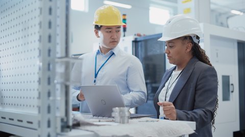 Two Professional Diverse Multiethnic Heavy Industry Specialist in Hard Hats Using a Laptop Computer at a Factory. Discussing an Industrial Machine Part, Comparing the Piece to Drafted Blueprints.