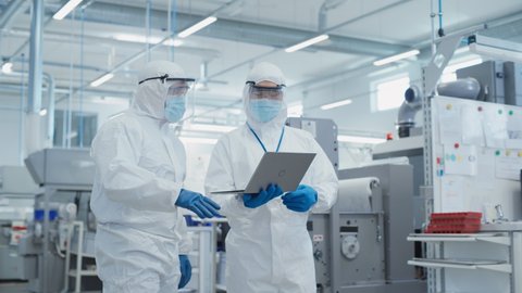 Two Scientists Standing in a Heavy Industry Factory in Sterile Coveralls and Face Masks, Using Laptop Computer. Examining Industrial Machine Settings and Configuring Production Functionality.