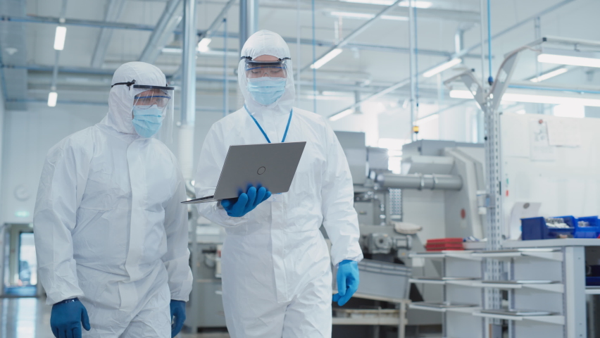 Two Scientists Walking in a Heavy Industry Factory in Sterile Coveralls and Face Masks, Using Laptop Computer. Examining Industrial Machine Settings and Configuring Production Functionality.