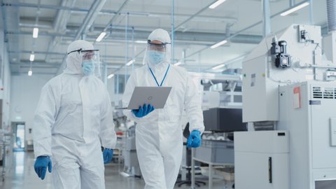Two Scientists Walking in a Heavy Industry Factory in Sterile Coveralls and Face Masks, Using Laptop Computer. Examining Industrial Machine Settings and Configuring Production Functionality.