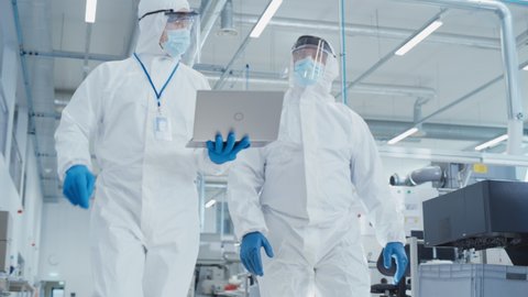 Two Scientists in Sterile Coveralls Walking in a Heavy Industry Factory with Laptop Computer, Examining Industrial CNC Machine Settings and Configuring Production Functionality.