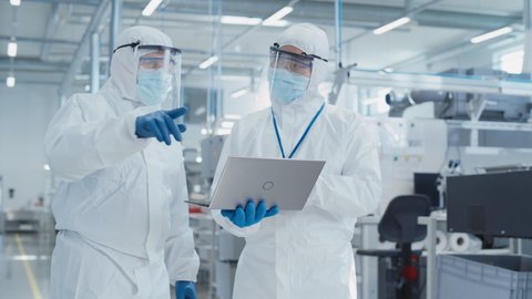 Two Engineers in Sterile Coveralls Standing in a Heavy Industry Factory, Using Laptop Computer, Examining Industrial Manufacturing Machine Settings and Configuring Production Functionality.