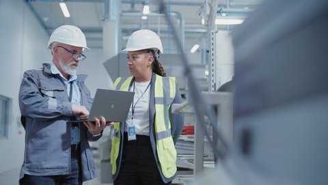 Portrait of Two Heavy Industry Employees in Hard Hats at Factory. Checking and Discussing Industrial Facility, Using Laptop Computer. African American Engineer and Middle Aged Technician at Work.