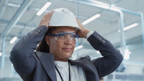 Close Up Portrait of a Middle Aged, Happy Black Female Engineer Putting On a White Hard Hat, While Standing at Electronics Manufacturing Factory. Heavy Industry Specialist Posing for Camera.