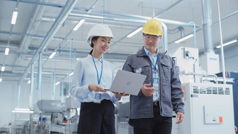 Two Young Asian Heavy Industry Engineers in Hard Hats Walking with Laptop Computer and Talking in a Factory. Two Manufacturing Employees at Work in Research and Development Facility.