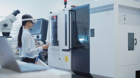 Female Engineer Wearing a Virtual Reality Headset and Operating a Heavy Industry Machine with Controllers at a Factory. Technician Configuring Industrial Machinery with the Help of VR Technology.