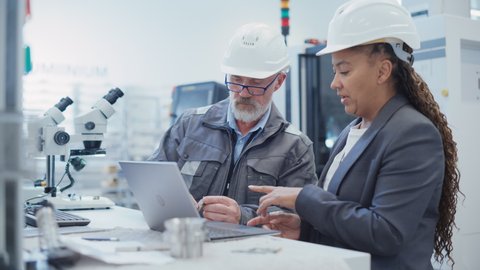 Two Professional Heavy Industry Employees Wearing Hard Hats at Factory. Checking and Discussing Industrial Machine Part, Working on Laptop Computer. African American Engineer and Technician at Work.