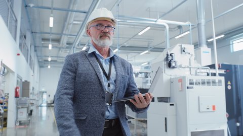 Portrait of a Bearded Middle Aged General Manager Walking in a Factory Facility, Wearing Casual Suit and a White Hard Hat. Heavy Industry Specialist Working on Laptop Computer.
