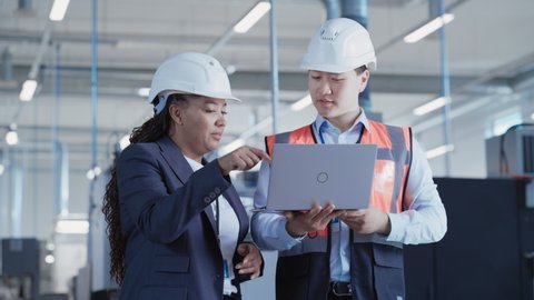 Professional Heavy Industry Employees Wearing Hard Hats at Factory. Checking and Discussing Industrial Facility, Working on Laptop Computer. African American Engineer and Asian Technician at Work.