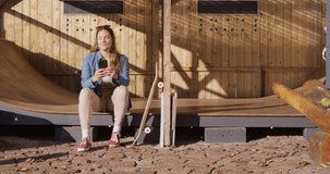 Video of happy caucasian female skateboarder resting and using smartphone. Skateboarding, sport, active lifestyle and hobby concept.