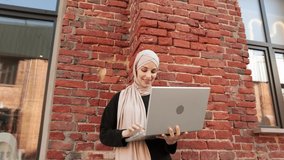 Pretty smiling muslim business woman in hijab standing near office centre and using laptop during video chat. Happy arabic girl standing outdoor near work office and checking email on digital gadget.