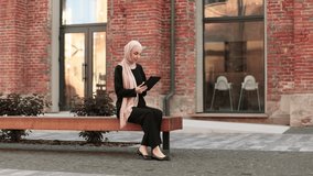 Portrait of smiling happy muslim woman relaxing using digital tablet while sitting on bench. Young muslim girl looking at screen of tablet speaking during online video call on social media on city.