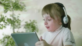 Funny Curious Child Little Girl Using Digital Tablet Technology Device Sitting At The Table Singing a Song Listening To Music Surfing Internet Play Game At Home