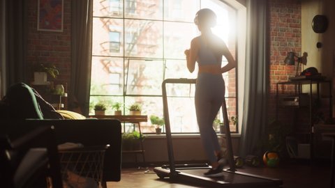 Beautiful Athletic Sports Woman Running on a Treadmill at Her Home Gym. Energetic Fit Female Athlete Training while Listening Podcast, Music in Headphones. Apartment with Window. Static Side Back View