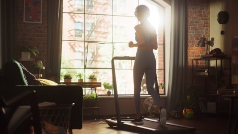 Beautiful Athletic Sports Woman Running on a Treadmill at Her Home Gym. Energetic Fit Female Athlete Training while Listening Podcast, Music in Headphones. Apartment with Window. Static Side Back View