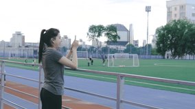 Young pretty female taking video of football players students playing football on the field at stadium. College student sportwoman doing live stream for sport supporters spectators