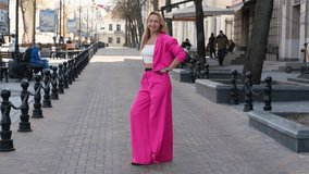 An adult woman in a bright pink pantsuit and a white top dances on a city street. Girl in a good mood. Slow motion video