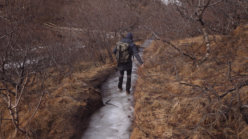 Back View of a Man Tourist with a Backpack Walking on a Slippery Ice Covered Trail Through Dry Spring Forest