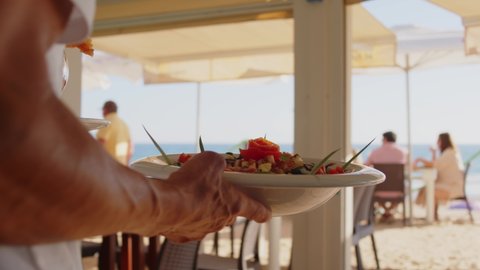 4K SlowMo - professional waiter bringing different plates - paella, salad, prawns in tempura - to a family's table in a restaurant on the beach : vidéo de stock